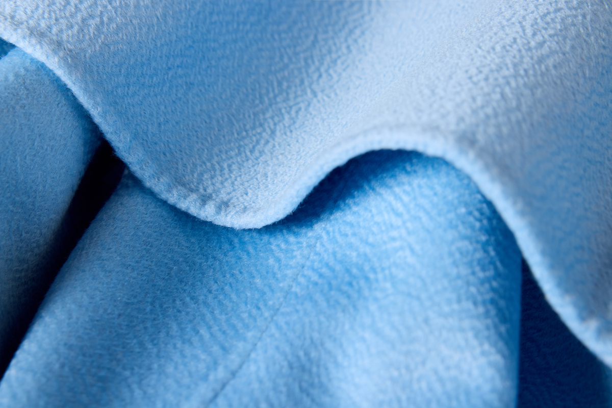 A close-up of light blue textured fabric, showing a soft woven surface with folded layers and a wavy edge.